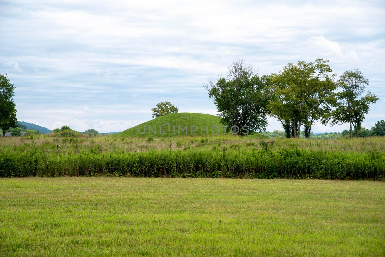 Grass covered prehistoric Hopewell culture burial mound at Siep Earthworks in Ohio under a dramatic cloudy sky with green grass and meadow foreground.