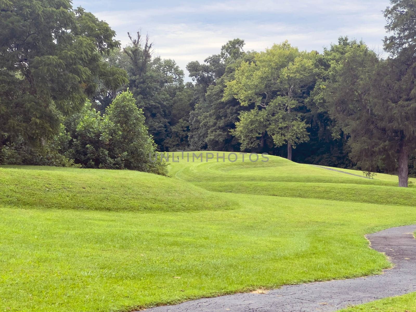 Walking path leads past curving body of a prehistoric Native American snake earthwork effigy mound at Great Serpent Mound in Ohio. Woodland background under a dramatic sky. Green grass is neatly trimmed and covers the curves of the snake. No people.
