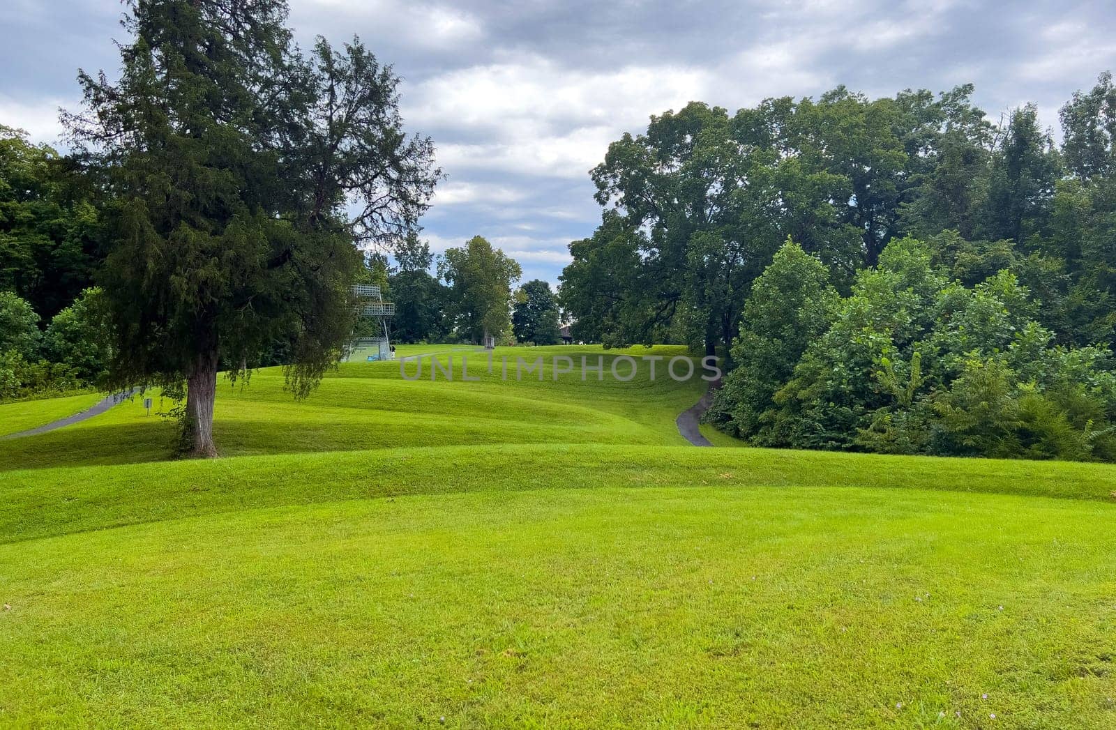 Curving body of a prehistoric Native American snake earthwork effigy mound at Great Serpent Mound in Ohio. Observation tower in background under a dramatic sky. Green grass is neatly trimmed and covers the curves of the snake. No people.