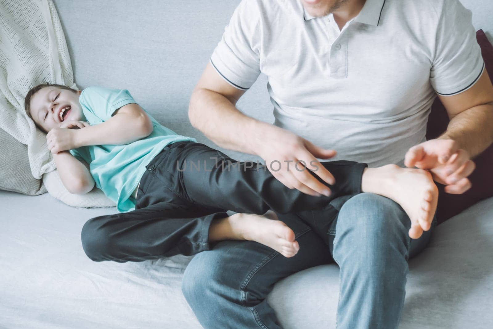 Father plays with his son 7-10 sitting on couch. Dad tickles kids feet. Family having fun
