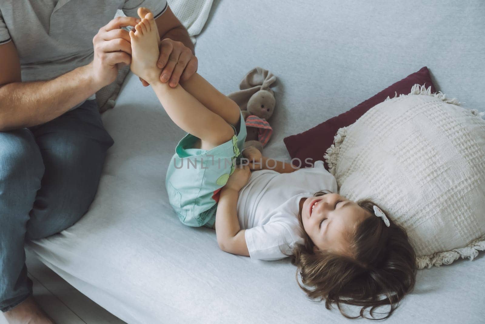 Father plays with little daughter 2-4 on couch. Dad tickles kids feet. Family having fun