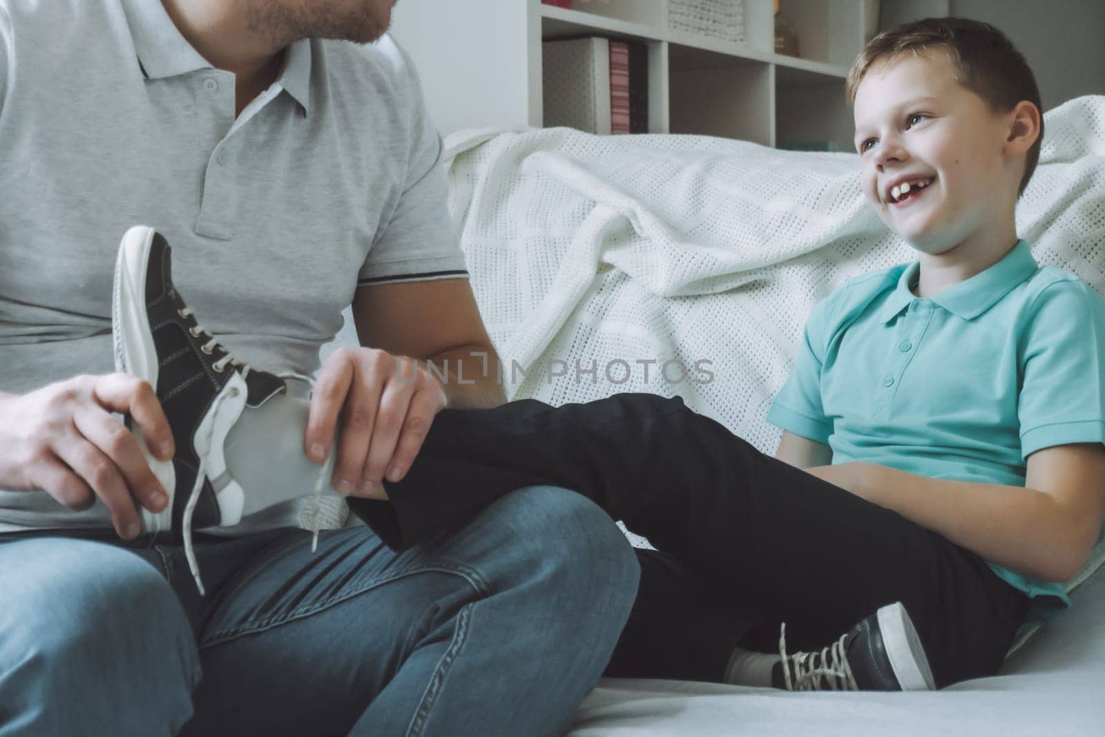 Dad and son sitting on couch. Father helps child to take off or put on shoes, kid laughs