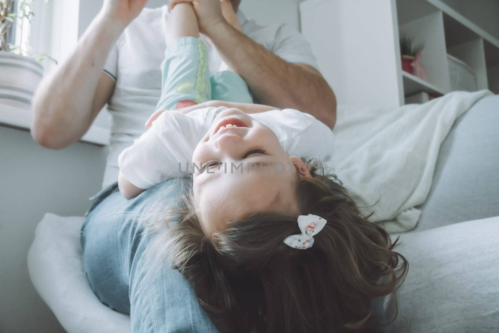 Dad and little girl 2-4 play on the couch. Kid lies on father's lap with her head down and laughs