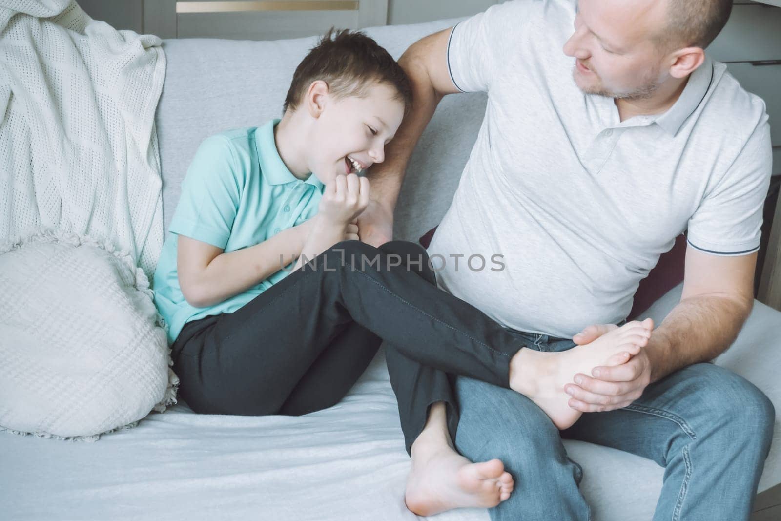Father plays with his son 7-10 sitting on couch. Dad tickles kids feet. Family having fun