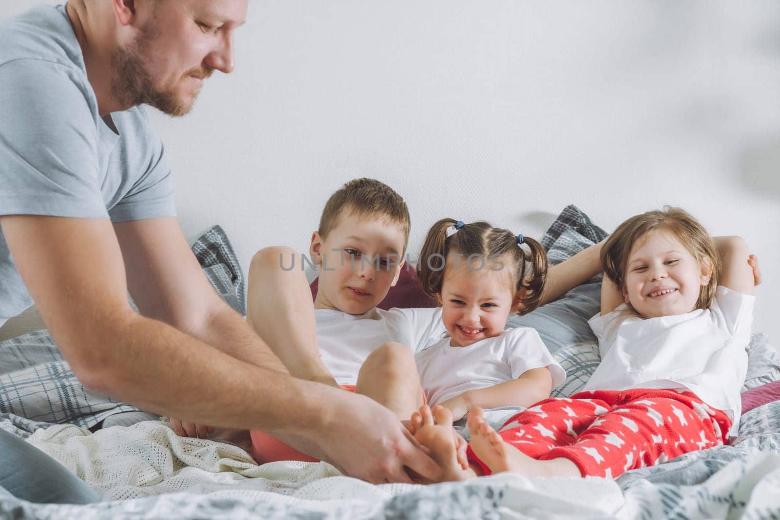 Father plays with three children on bed. Dad tickles kids feet. Family of daddy, two girls and boy