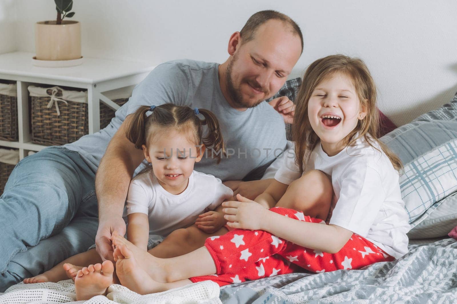Father plays with two children on bed. Dad tickles kids feet. Family of daddy and two little girls