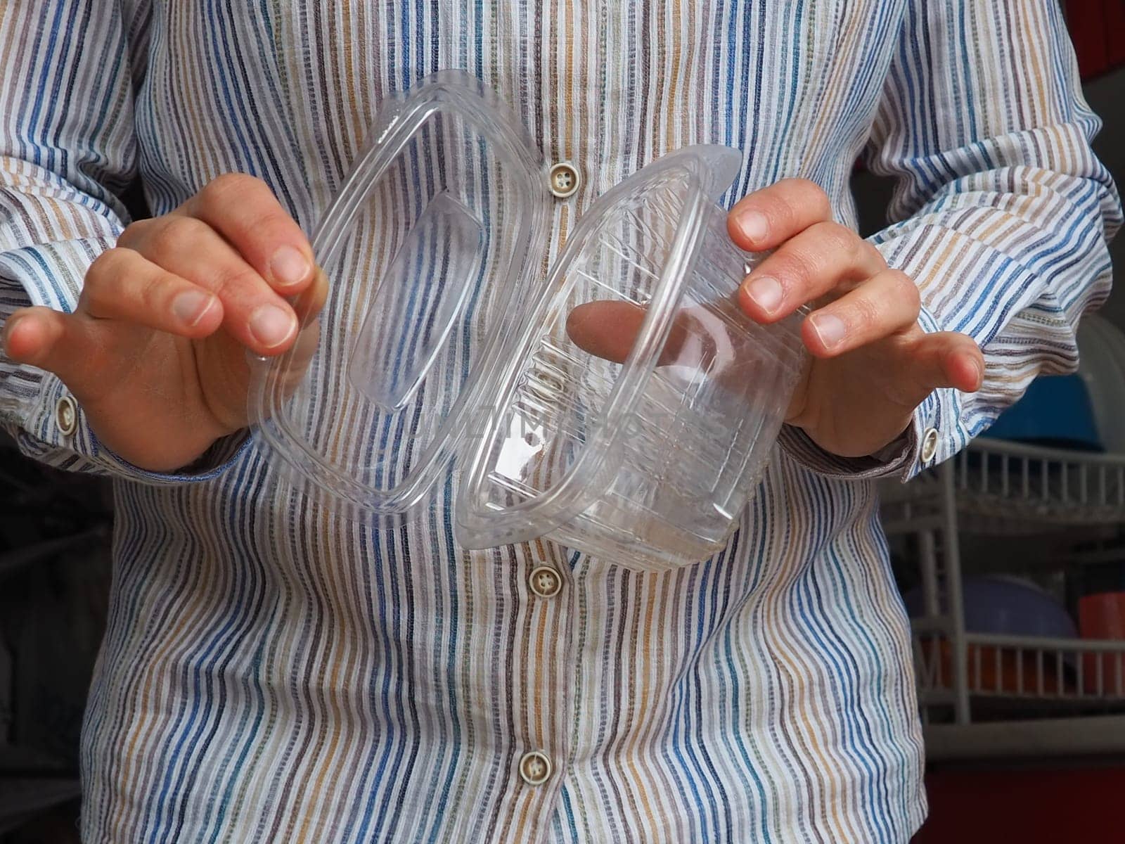 Female hands hold a plastic container or packaging. Kitchen utensils. A woman in a white striped shirt stands in the kitchen and shows a disposable plastic box with a lid. Hygiene and recycling theme.
