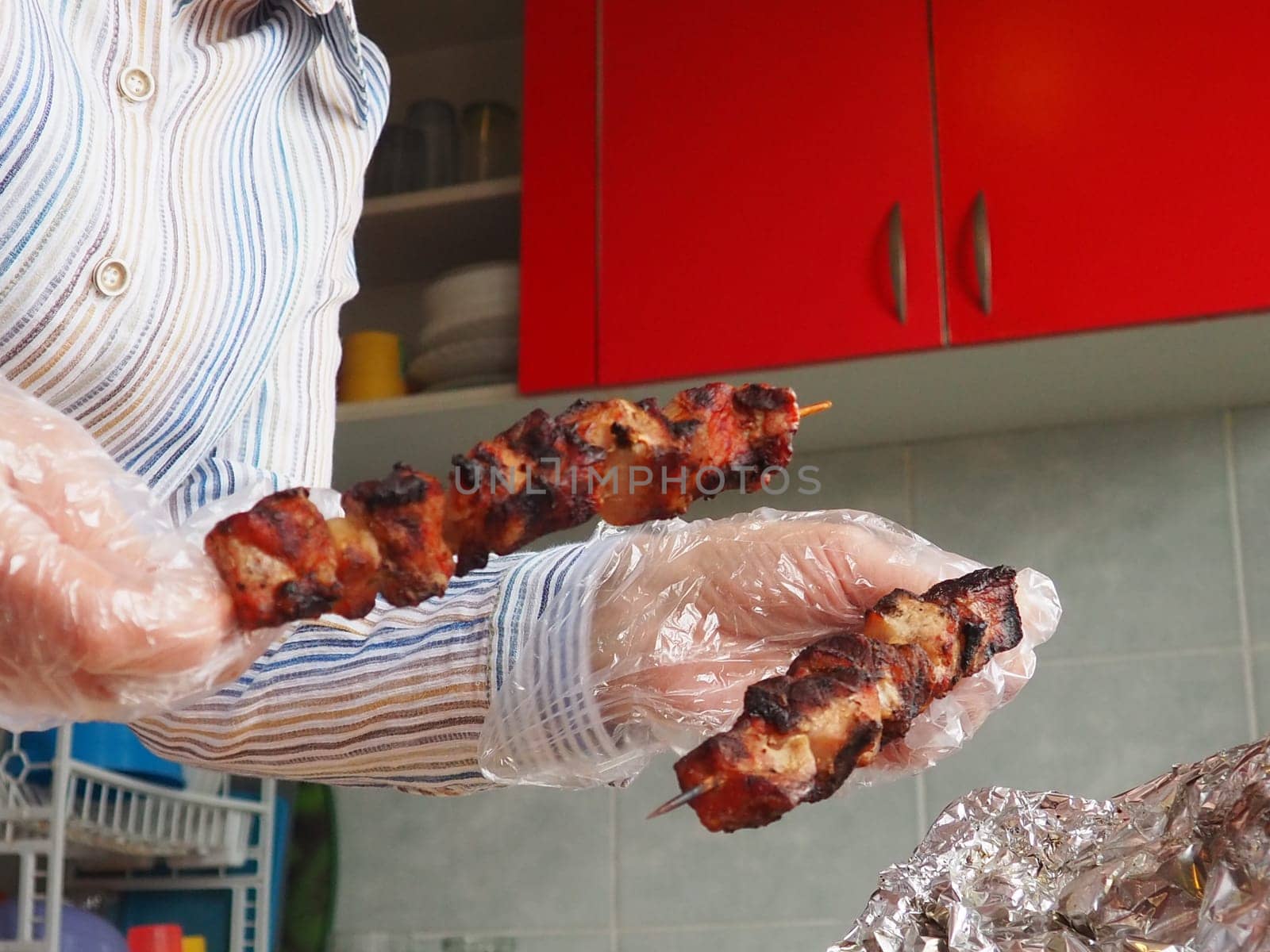 The grilled meat was delivered from the restaurant. Home delivery from restaurants. Women's hands in disposable gloves hold meat on a stick. Kitchen with red furniture. Cleanliness and safety of food.