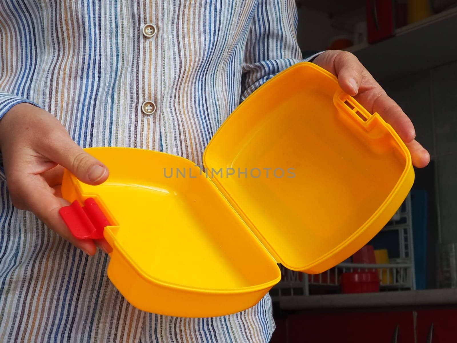 Female hands hold a yellow open plastic school lunch container or packaging. Kitchen utensils. A woman in a white striped shirt shows the lunch box. Food hygiene and recycling theme.