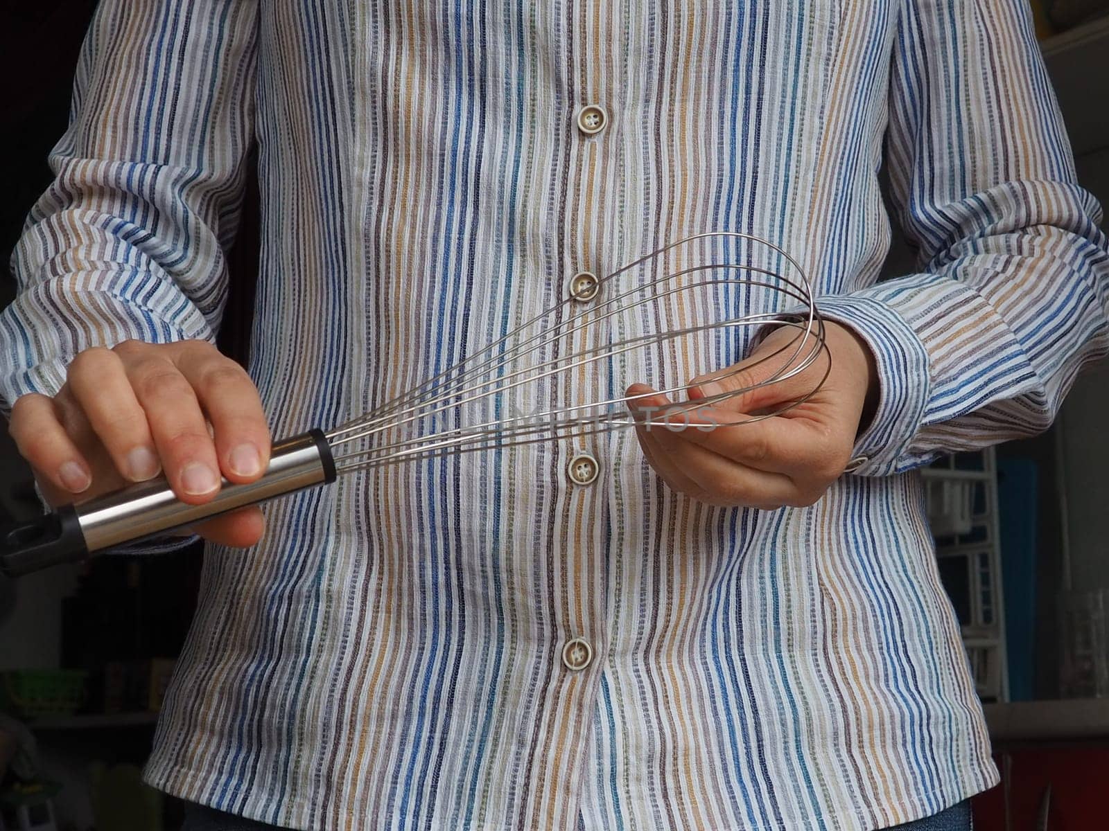 Women's hands hold a metal whisk for whipping or kneading. Kitchen and cooking utensils, dishes. A woman in a white striped shirt stands in the kitchen and shows a whisk.