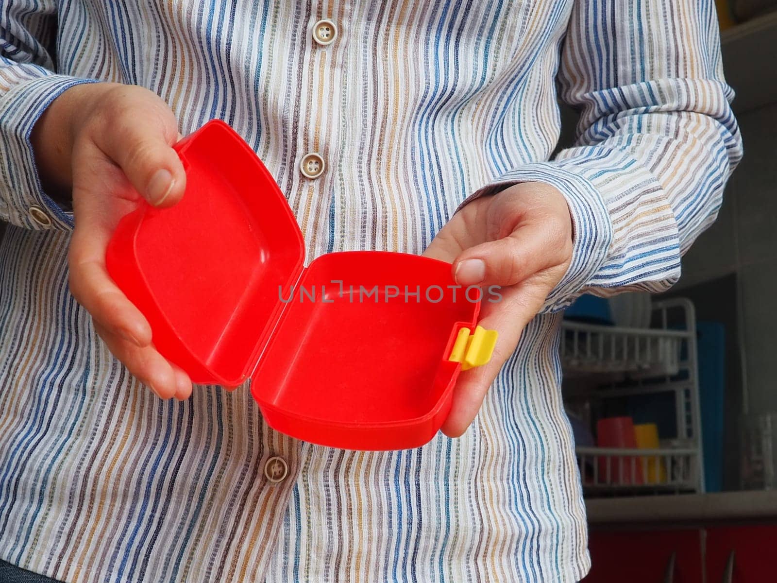 Female hands hold a red open plastic school lunch container or packaging. Kitchen utensils. A woman in a white striped shirt shows the lunch box. Food hygiene and recycling theme.