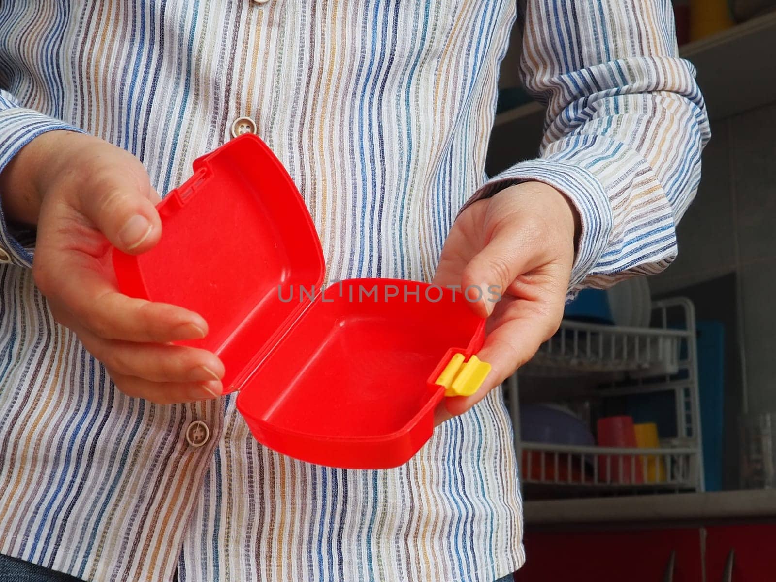 Female hands hold a red open plastic school lunch container or packaging. Kitchen utensils. A woman in a white striped shirt shows the lunch box. Food hygiene and recycling theme.