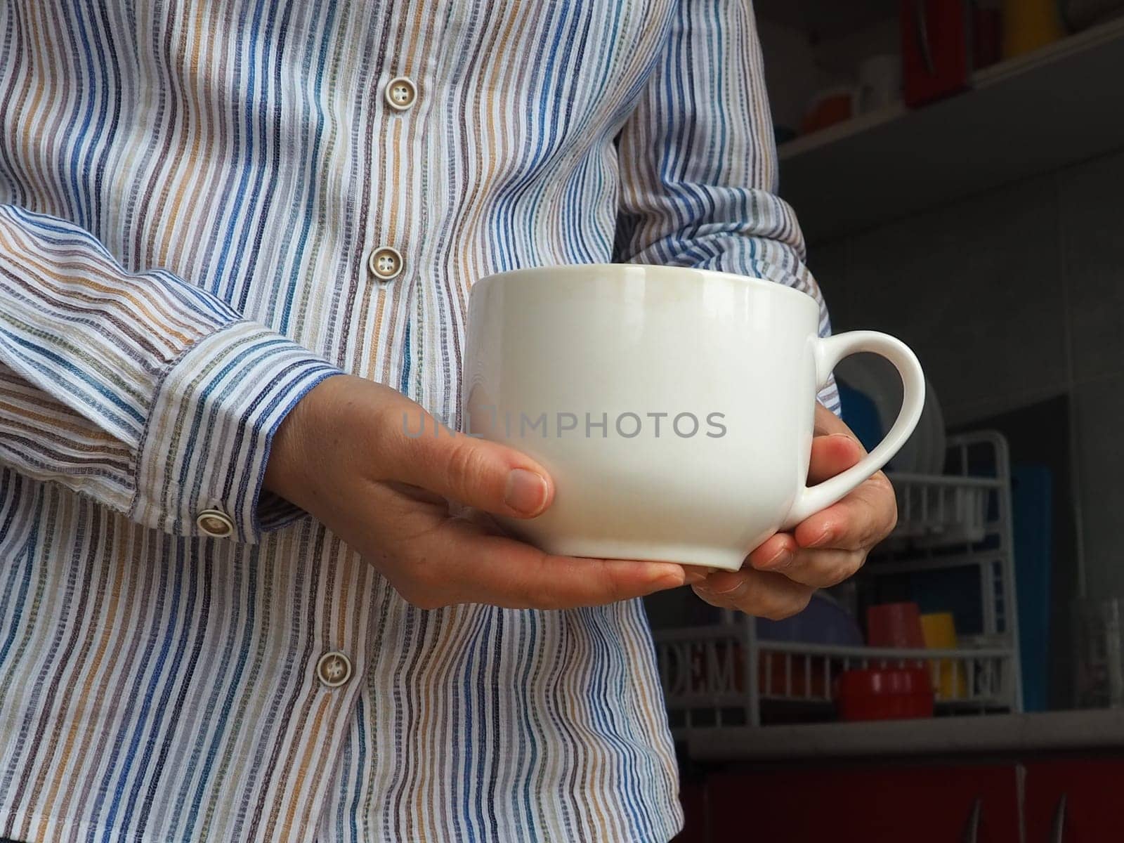 Female hands hold a large white ceramic mug or cup. Kitchen and cooking utensils, dishes. A woman in a white striped shirt stands in the kitchen and shows an unmarked cup. Copy space.