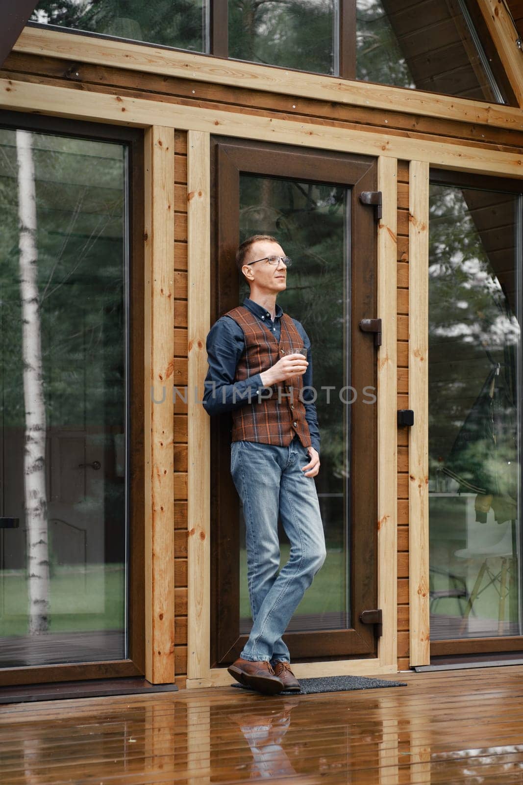 Pensive middle age man stands on the terrace of a wooden bungalow and drinks bourbon
