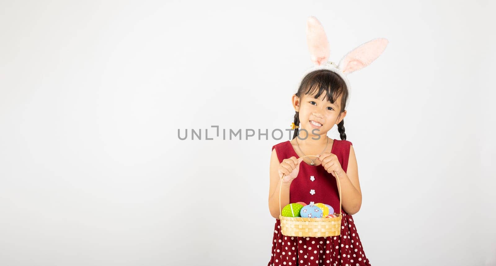 Happy Easter Day. Smile Asian little girl wearing easter bunny ears holding basket of full colorful eggs smiles broadly isolated on white background with copy space, Happy child in holiday