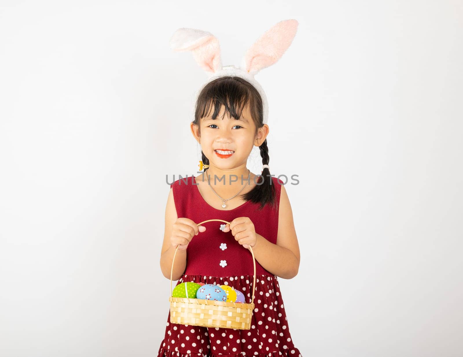 Happy Easter Day. Smile Asian little girl wearing easter bunny ears holding basket of full colorful eggs smiles broadly isolated on white background with copy space, Happy child in holiday