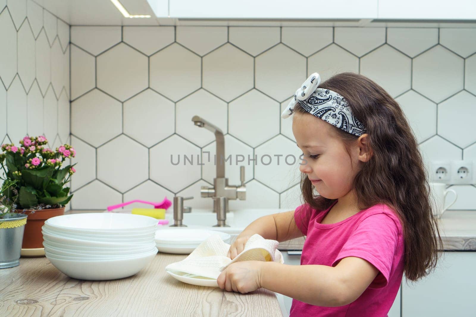 Happy little girl wiping clear plates with dry towel on kitchen set. Portrait of child washing up dishes in kitchen sink side view. Home cleaning concept. Dry tableware after washing.