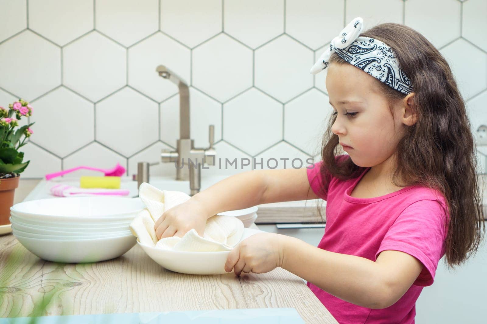Upset little girl wiping clear bowls with dry towel on kitchen set. Portrait of child washing up dishes in kitchen sink side view. Home cleaning concept. Dry tableware after washing.