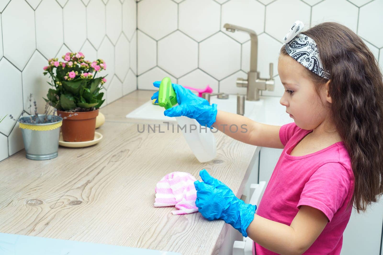 Nice little girl in protective gloves wipe kitchen set with household rag and detergent sprayer. Portrait of child tidying up kitchen side view. Home wet cleaning. Detergent and cleaning supplies.
