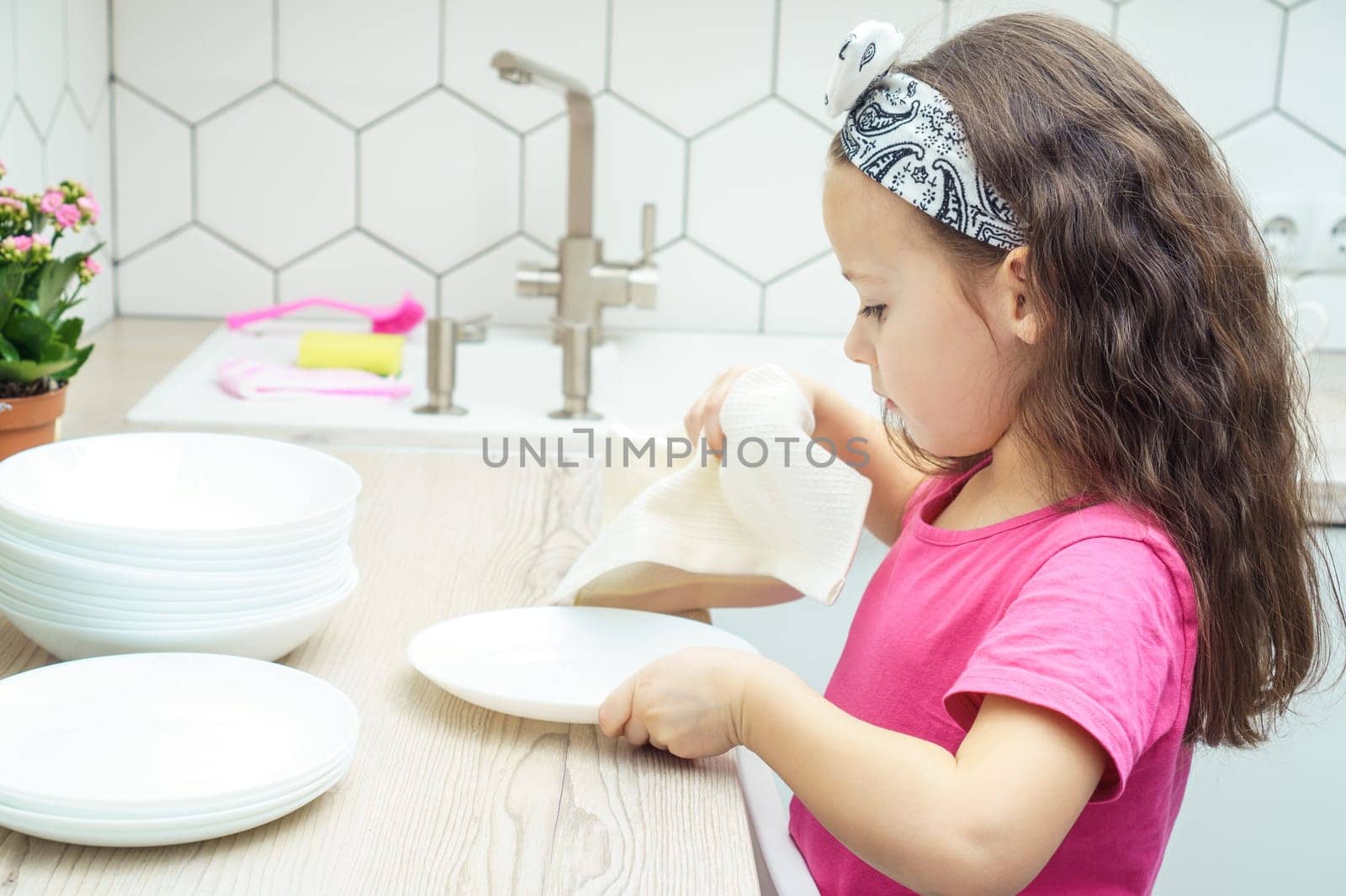 Pretty little girl wiping clear white plates with dry towel on kitchen set. Portrait of child washing up dishes in kitchen sink side view. Home cleaning concept. Dry tableware after washing.