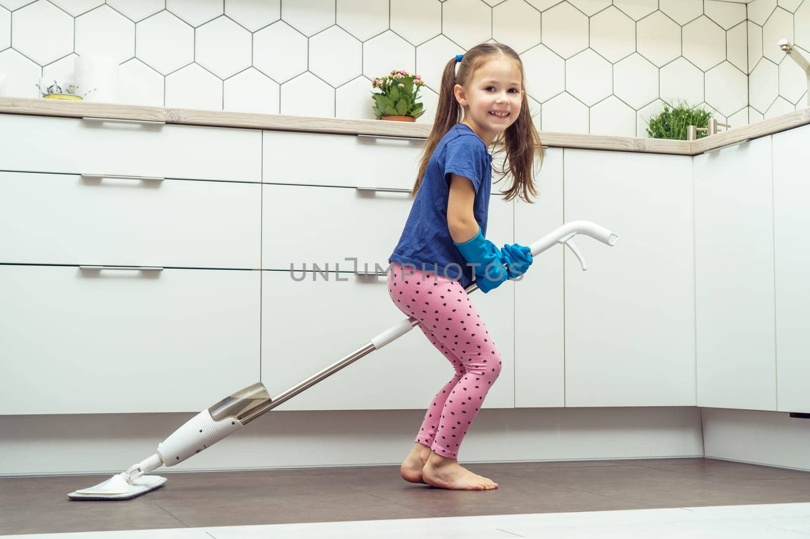 Happy little girl sitting on modern mop for floor washing with removable brush near kitchen set. Full length photo of child tidying up kitchen side view. Home cleaning concept. Kid help clean house.