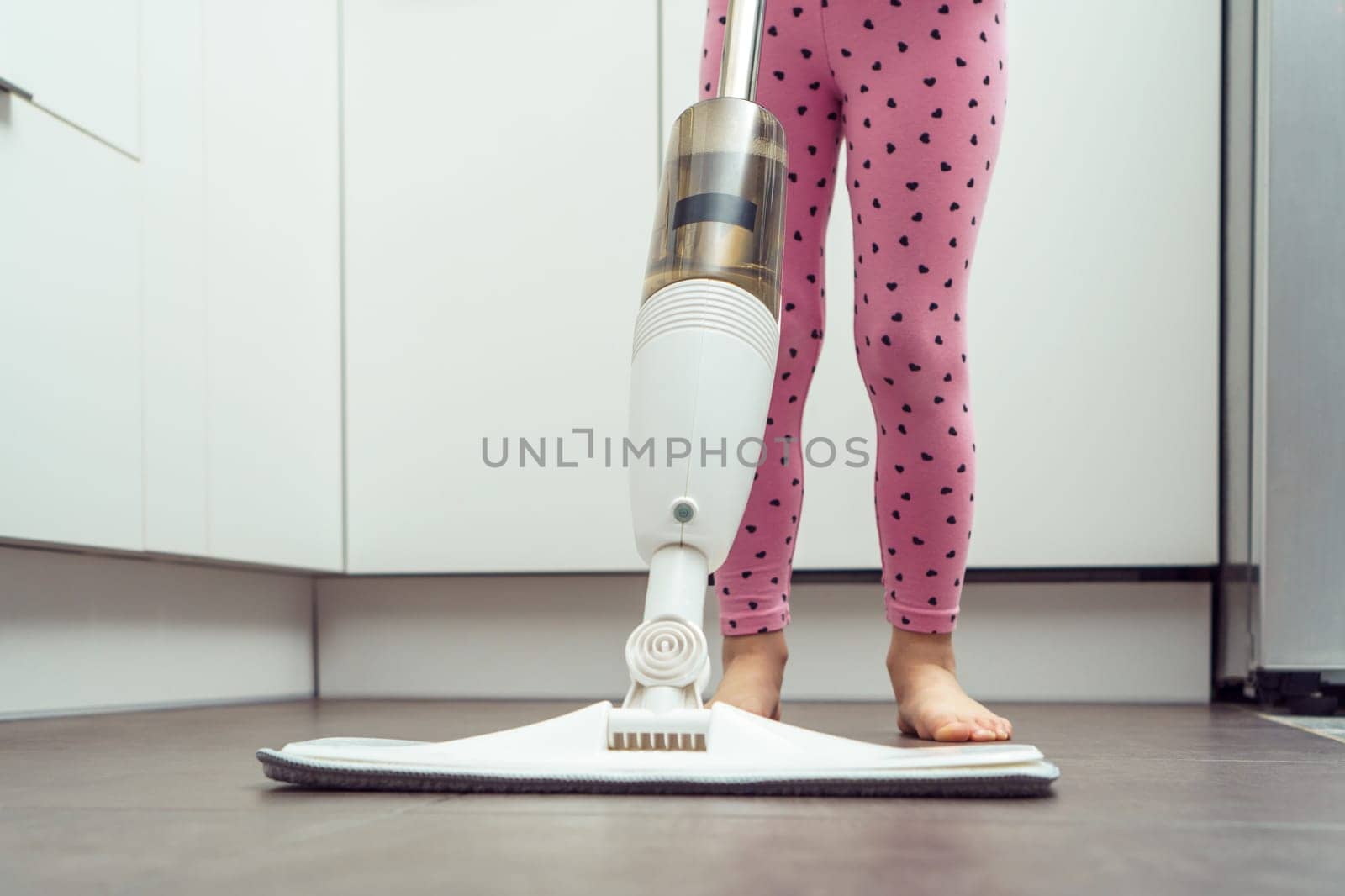 Little girl wash floor by modern mop with removable brush and water container near kitchen set, cropped photo closeup. Tidying up kitchen, home wet cleaning. Technologically advanced appliance.