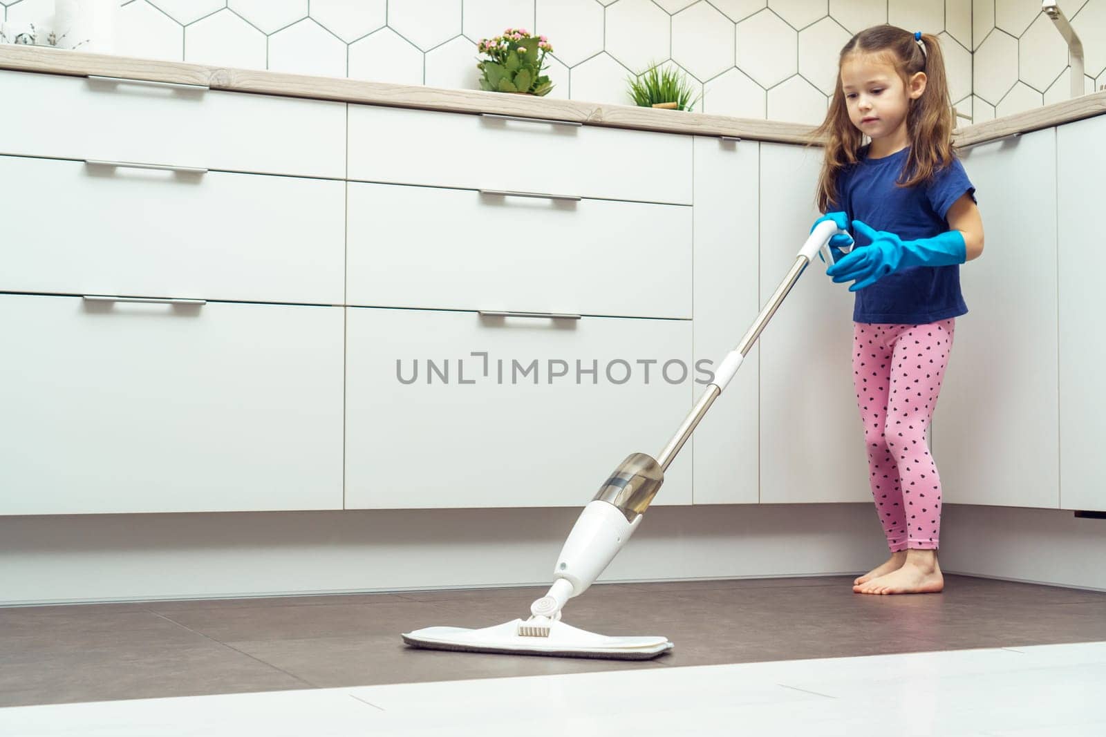 Pretty little girl in household gloves wash floor by modern mop with removable brush and water container near kitchen set. Full length photo of child tidying up kitchen side view. Home wet cleaning.