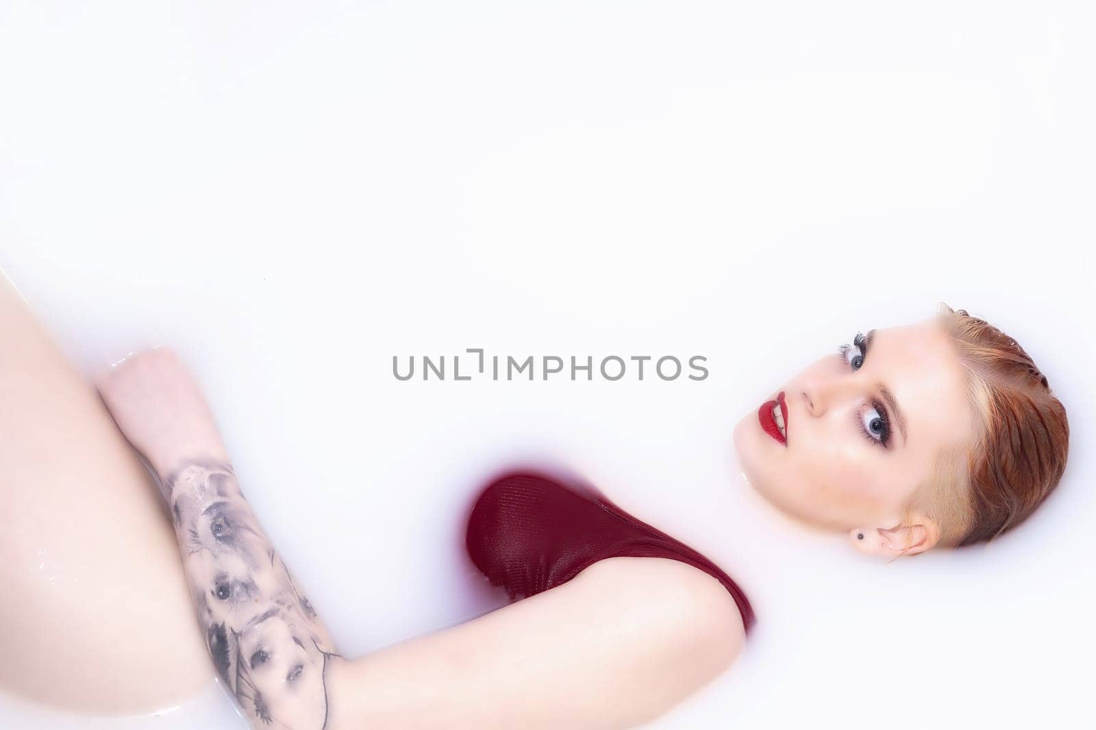 Attractive young woman with blue eyes, red lips and tattoo is lying in spa bath with milk and looking at camera. Horizontally.