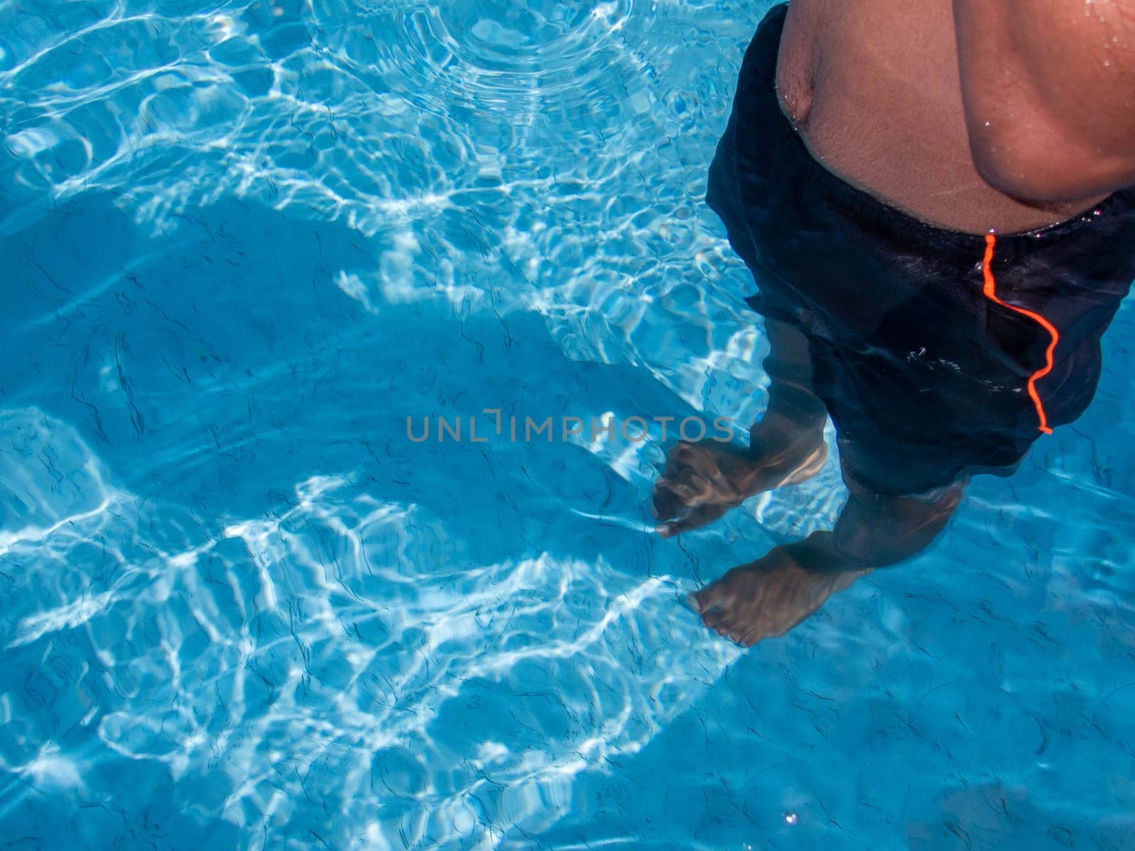 High angle view of torso and legs of a teenage boy standing in clear blue swimming pool water wearing swimwear. Shot in natural summer sunlight, his shadow is on the pool floor with waves and ripples. With copy space.