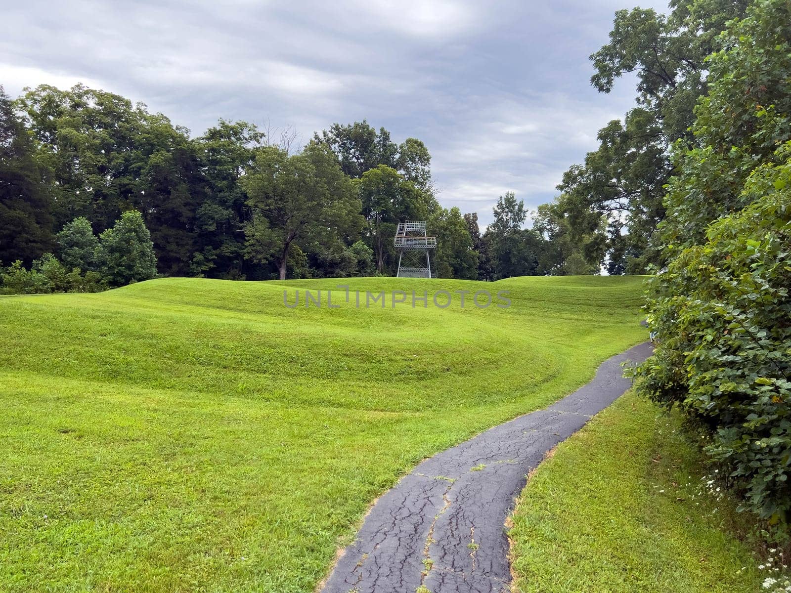 Walking path leads past curving body of a prehistoric Native American snake earthwork effigy mound at Great Serpent Mound in Ohio. Observation tower in background under a dramatic sky. Green grass is neatly trimmed and covers the curves of the snake. No people.