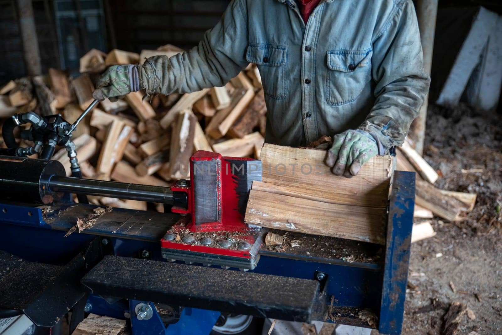 Work man splits wood with a hydraulic log splitter. Blade is seen cutting into the wood. Defocused wood pile in background