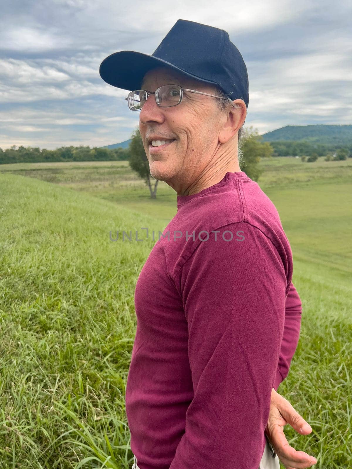 Caucasian senior man with glasses and a hat turns smiling toward camera on the top of a green grass covered hill under a dramatic sky. Active lifestyle and wellbeing concepts, as well as success and achievement.