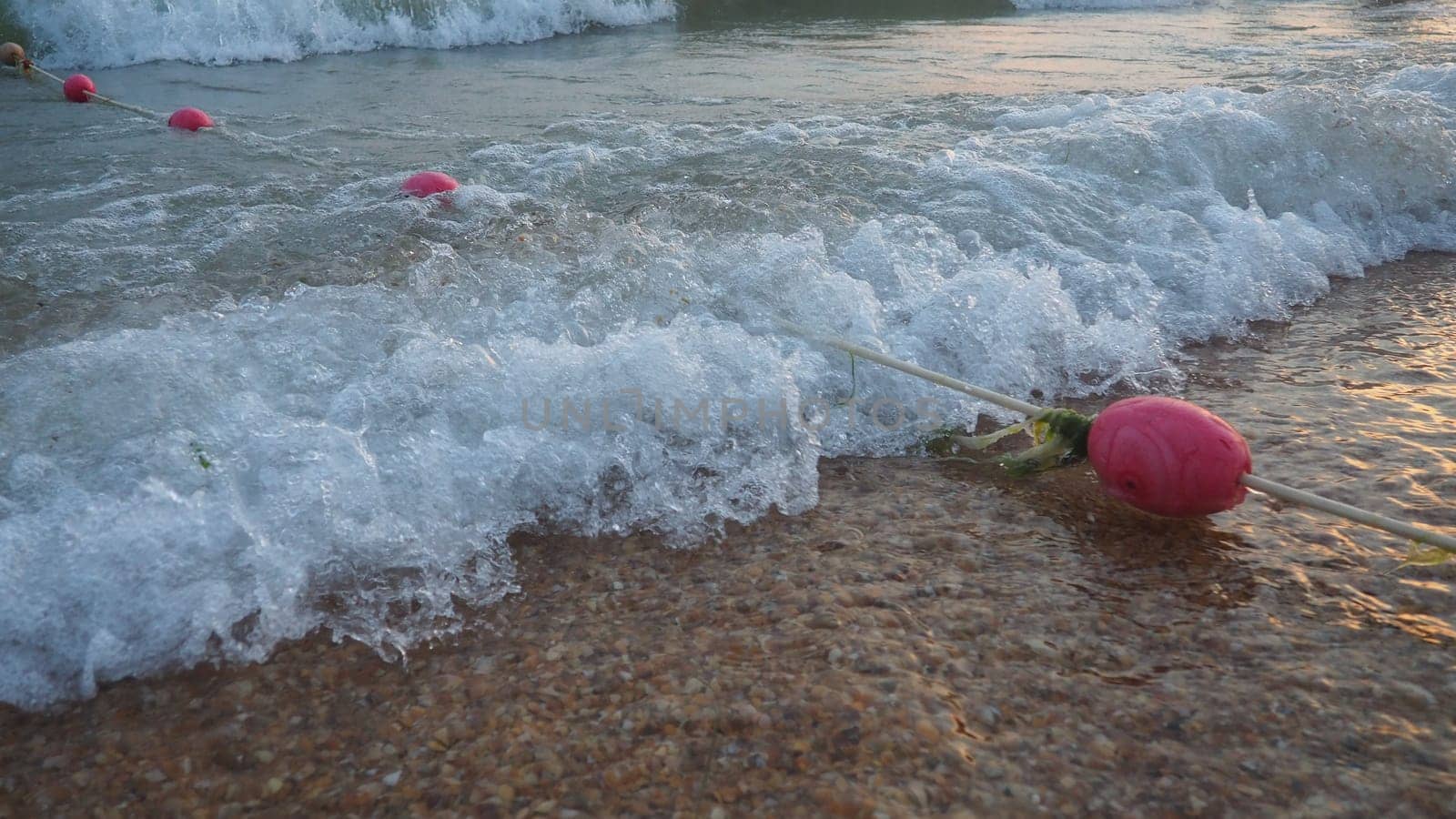 Buoys on a rope near sea water. The buoys are pink restraints to alert people to the depth of the water. Rescue of the drowning. Delimiting a place on a sandy beach between hotels. Wave with bubbles