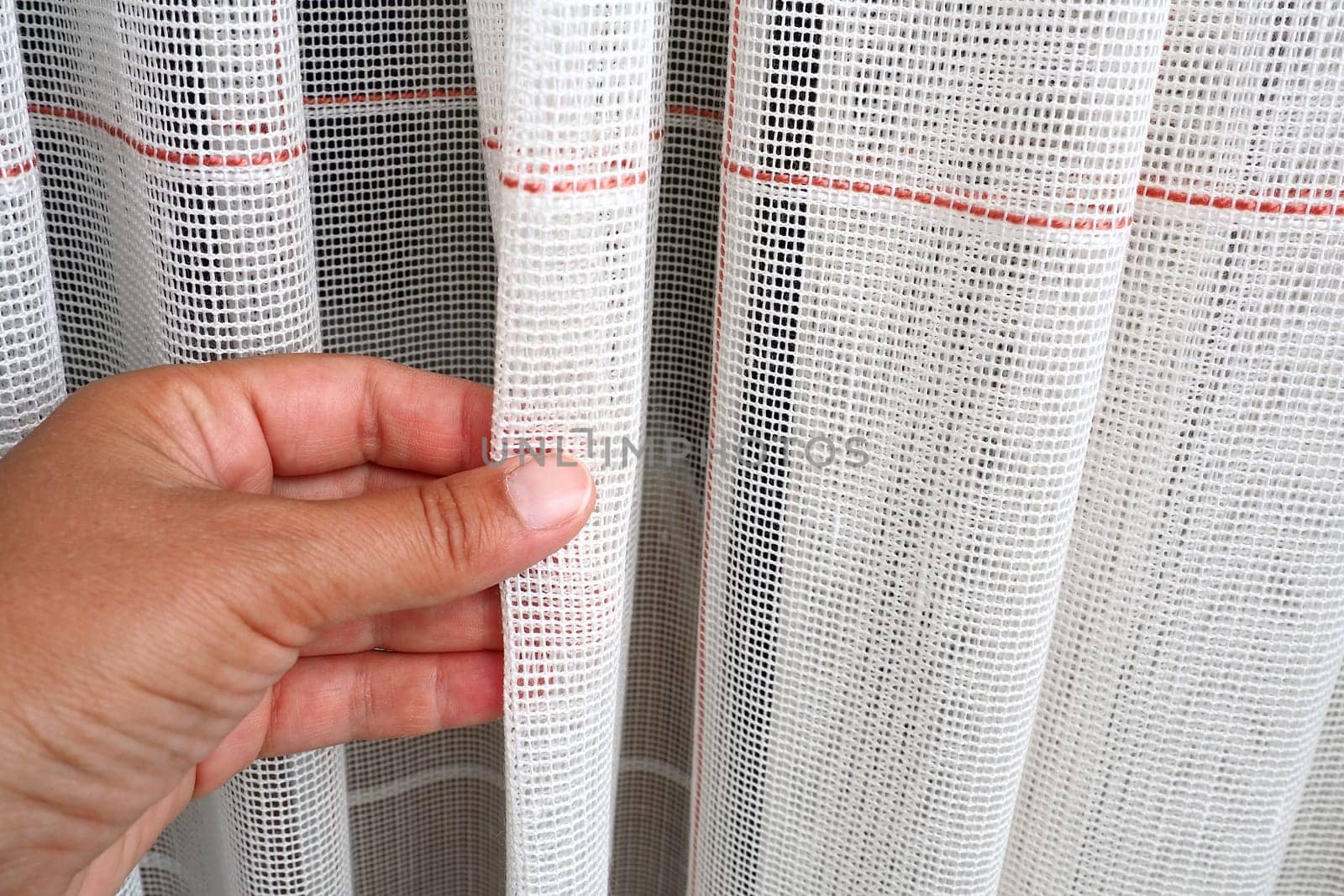White coarse cotton or linen curtains made of weaves of thin threads. Simple compact economical apartment interior. Window decoration in the room. A woman's hand strokes the tulle, shows the quality