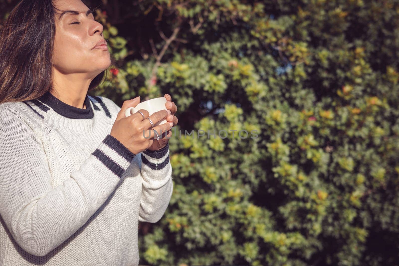 A Latina woman sits on a wooden bench in the garden, enjoying the fresh air while taking some time for herself. She holds a cup of coffee and smiles with a relaxed expression, enjoying a moment of tranquility.