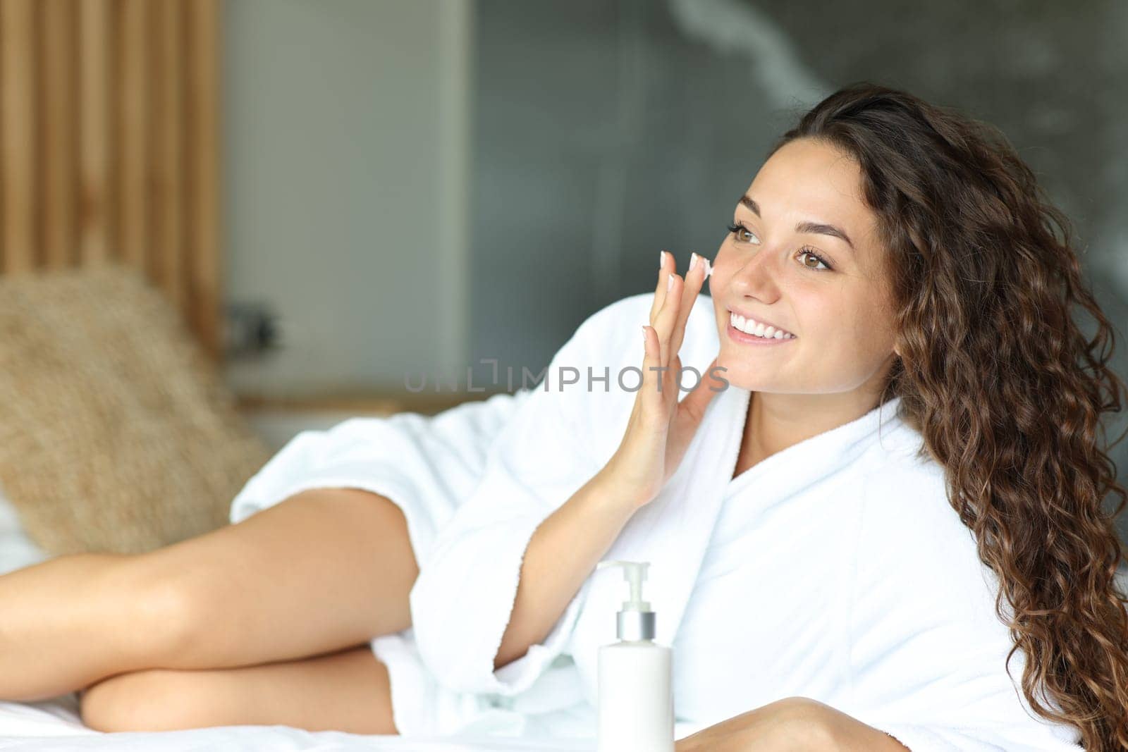 Happy woman hydrating face skin applying moisturizer on a bed at home