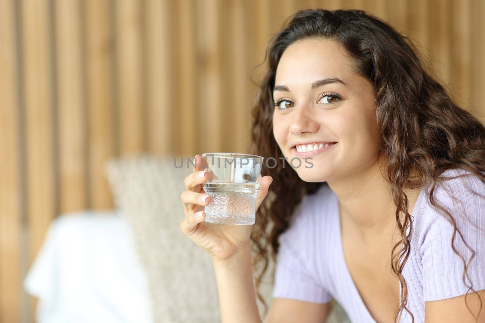 Happy woman holding water glass looks at you sitting on a bed