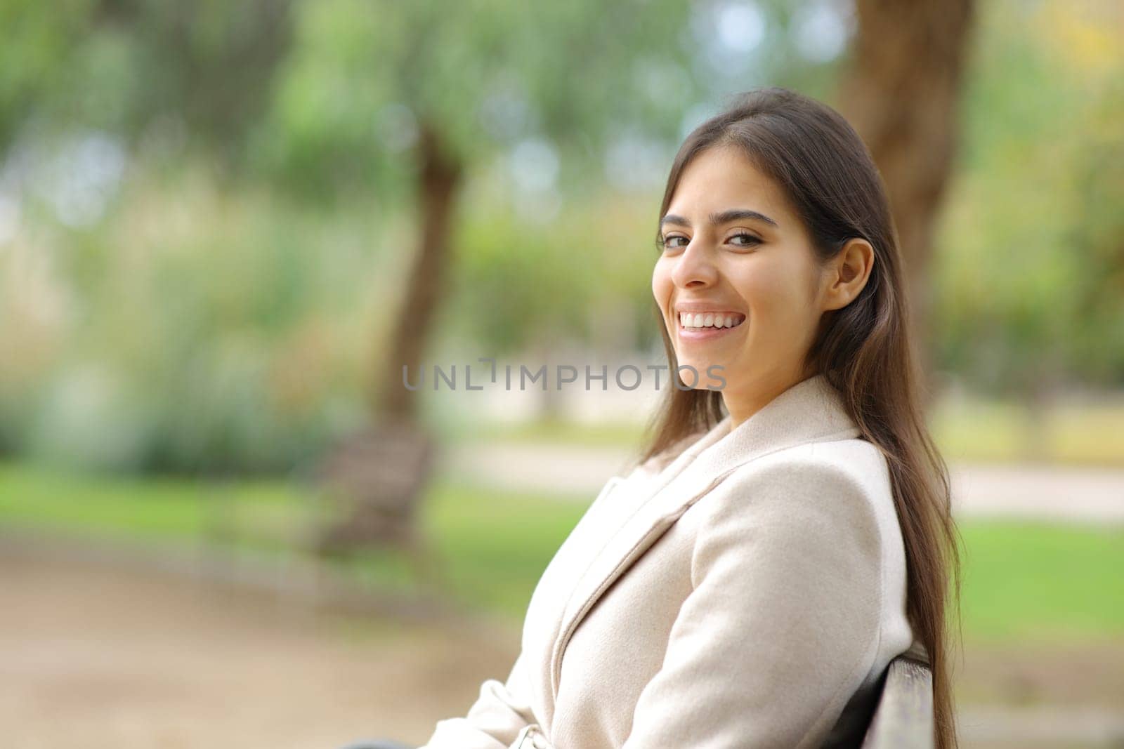 Happy woman in winter looking at camera sitting on a bench in a park