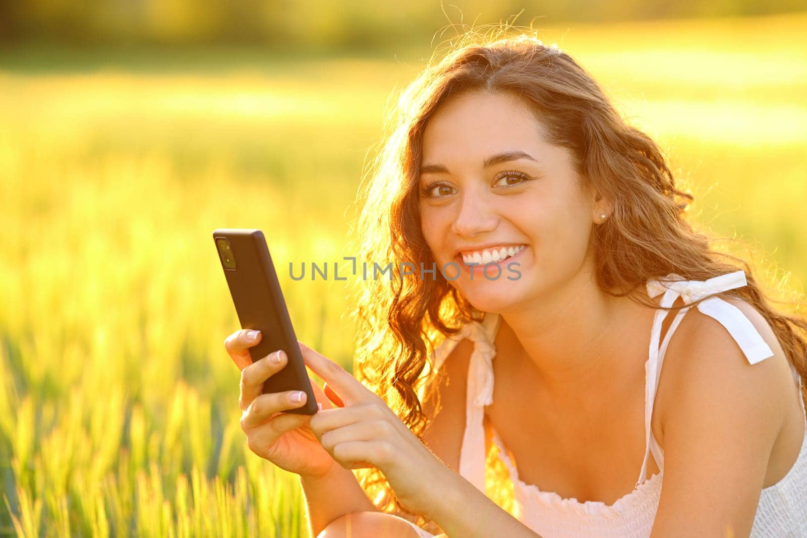 Happy woman posing in a field holding cell phone at sunset
