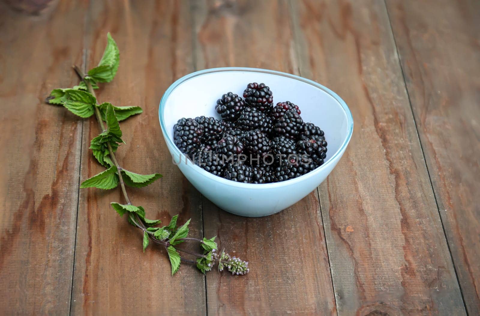 In a glass bowl are many ripe blackberries. Next to them a sprig of mint.