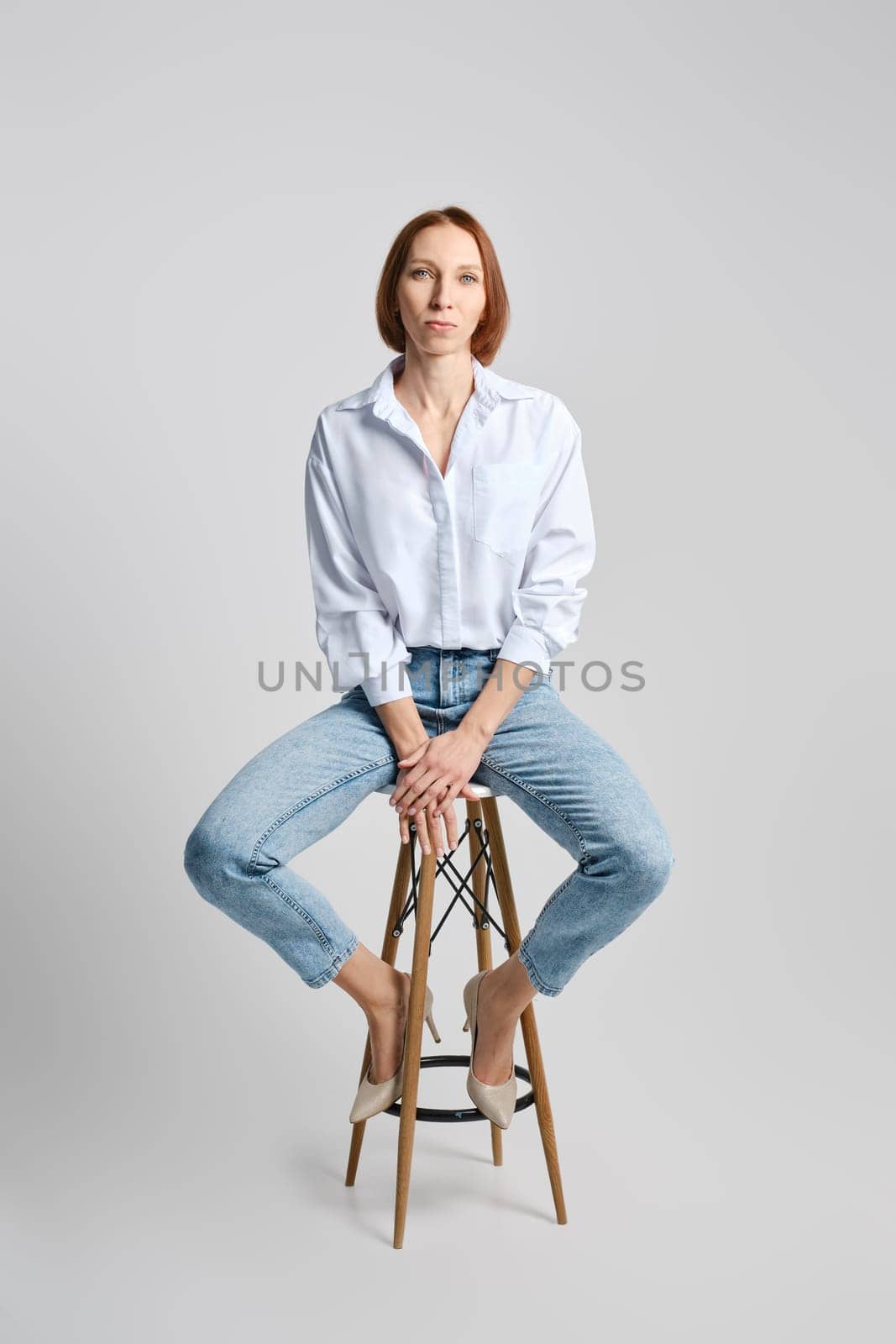 Lifestyle portrait of middle aged caucasian redhead woman in white shirt and blue jeans sitting on tall stool