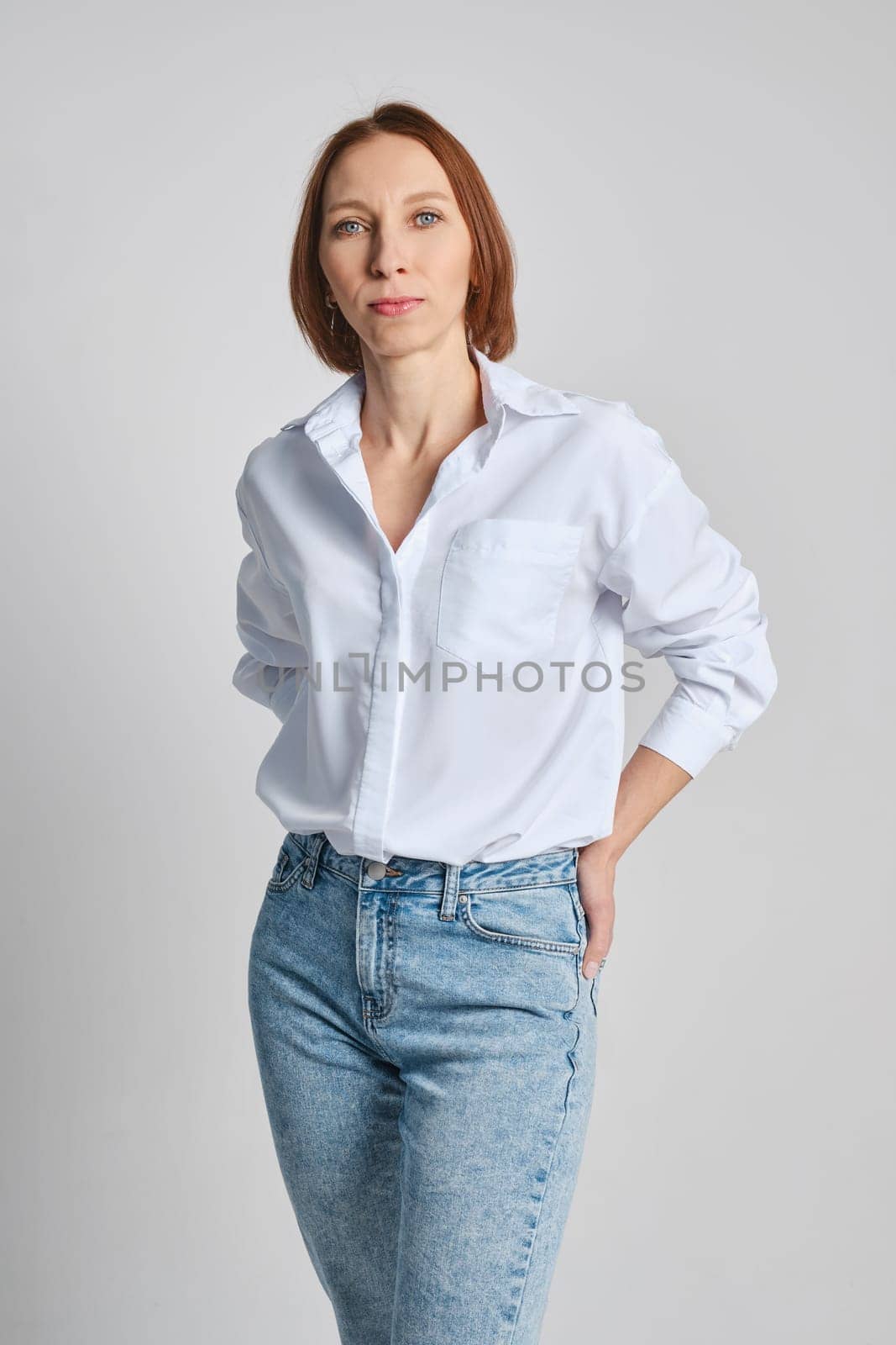 Portrait of a confident stylish redhead woman standing against grey background with arms in back pocket of her jeans