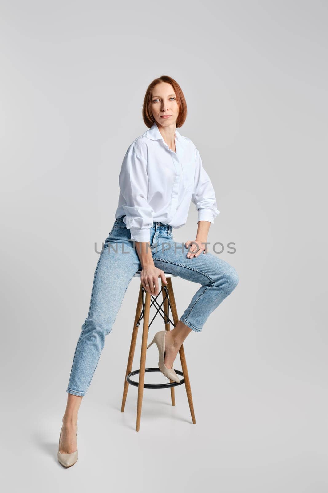 Casual portrait of middle aged redhead woman in white shirt and blue jeans sitting on tall stool and stretching one leg to the side