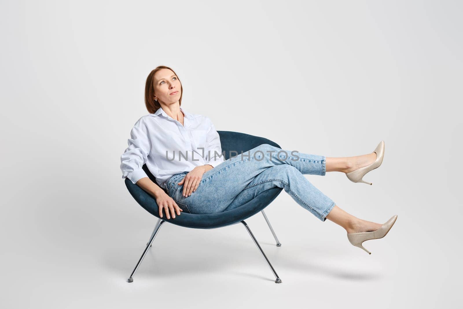 Carefree middle aged woman sitting in cosy armchair and waiving her long legs
