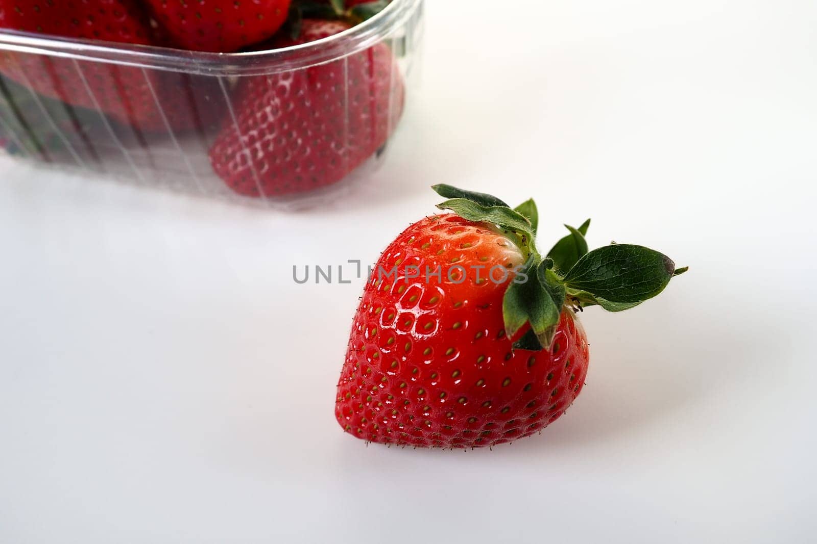 Ripe strawberries in a plastic package on a white background. Delicious fresh berries in a container for sale to customers. Keeping food fresh. Healthy food. One strawberry pulled out of the container