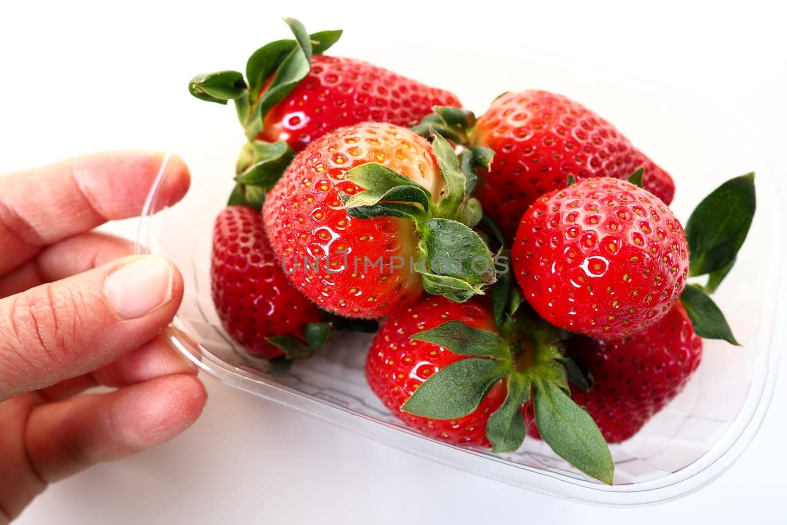 Ripe strawberries in a plastic package on a white background. Delicious fresh berries in a container for sale to customers. Keeping food fresh. Healthy food. Woman's hand holding strawberries