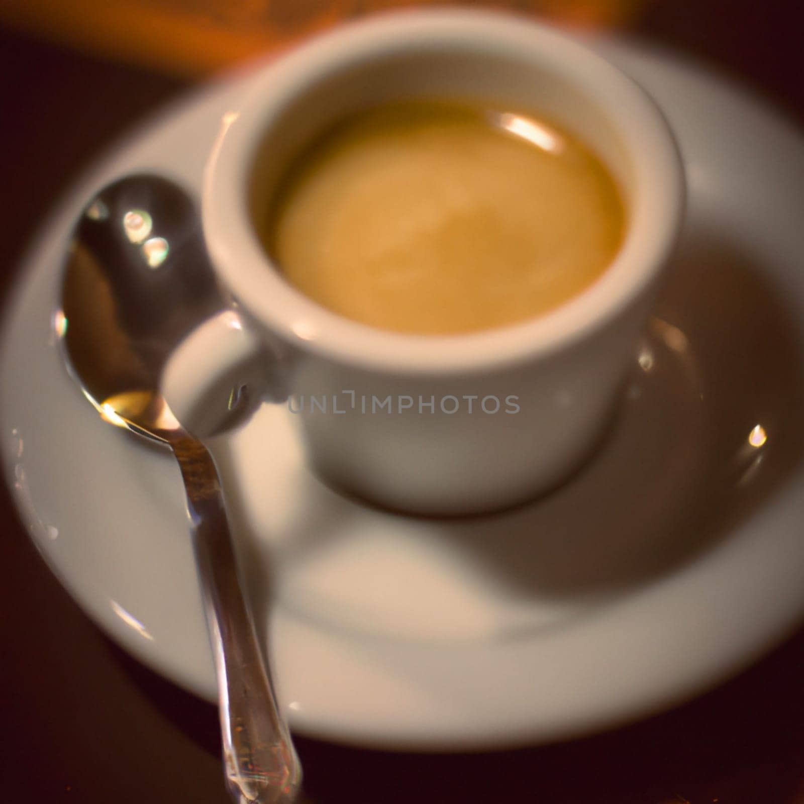 Cup of coffee in cafe. Anamorphic bokeh effect. Tilt-shift photo. Hot coffee in a white cup with a saucer and a spoon. The coffee is getting cold. Aroma of coffee beans. Label or advertisement. Foam