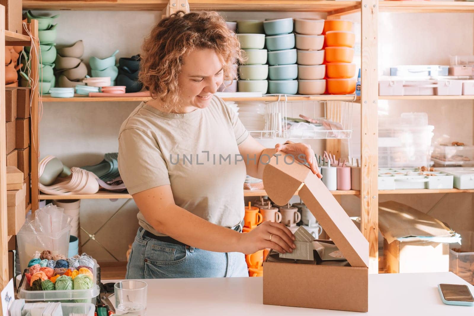 Smiling blond curly woman packing and wrapping shipment with postcards, silicone baby dishes in cardboard delivery box. Colorful skein of thread in container on desk. Workshop store, small business
