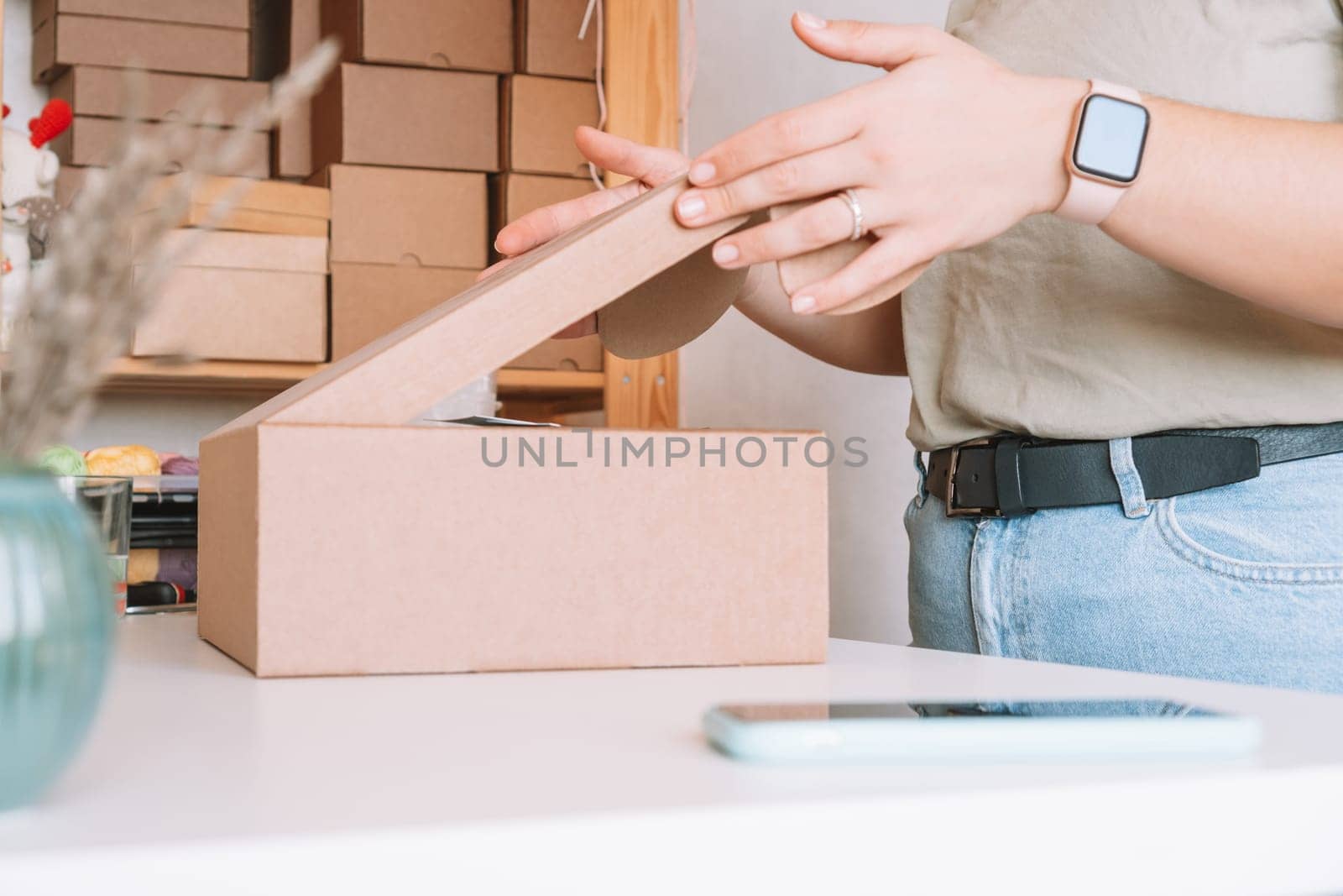 Cropped photo of woman hands packing, preparing parcel for delivery package to customers. Small business owner, individual entrepreneur in workshop, startup. Mobile phone on desk. Online order