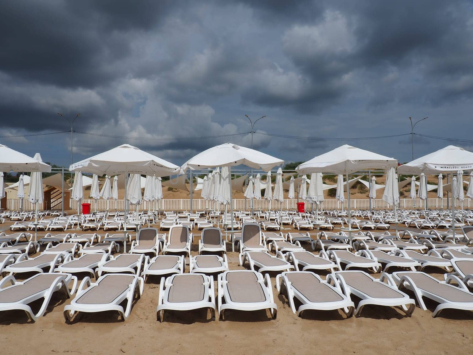 Wet sunbeds and umbrellas on the sea beach during heavy rain storm. Rolled up parasol on the beach against dark cloudy sky. Invasion of a typhoon, cyclone or storm on the sea or ocean. Beach season
